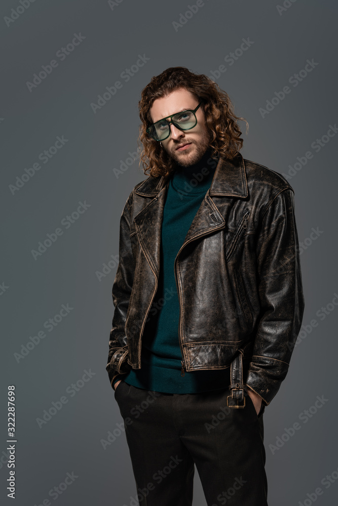 handsome man in leather jacket with hands in pockets looking at camera isolated on grey