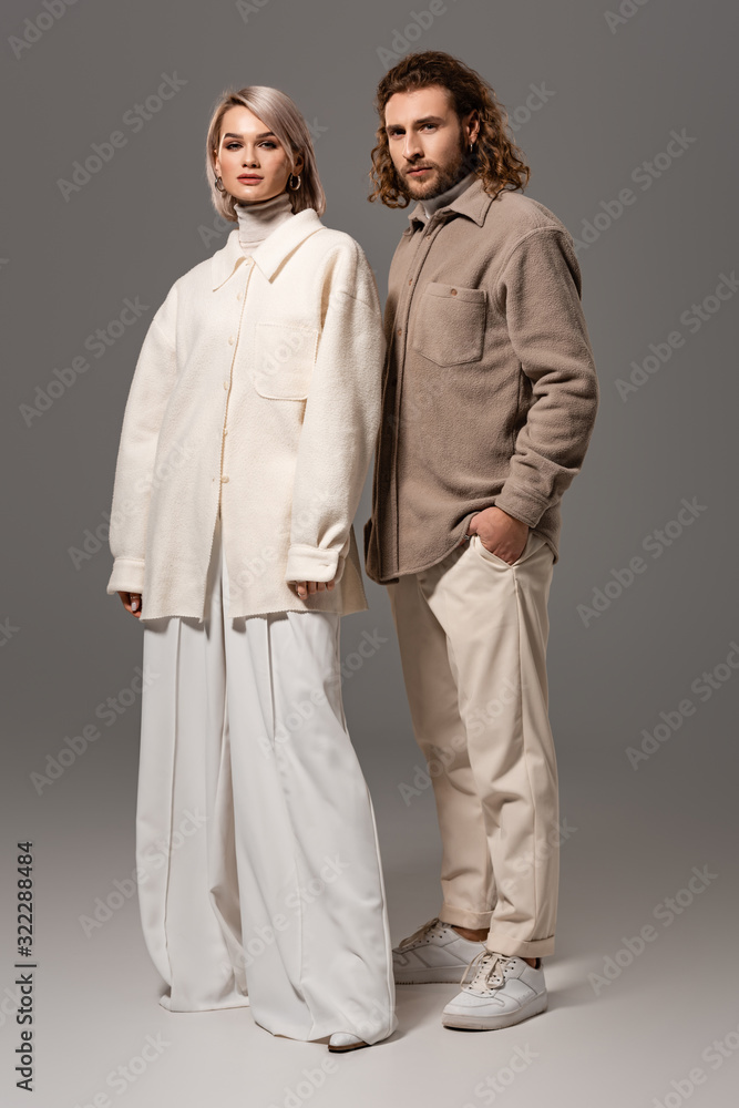 woman in white coat and man in shirt with hand in pocket looking at camera on grey background