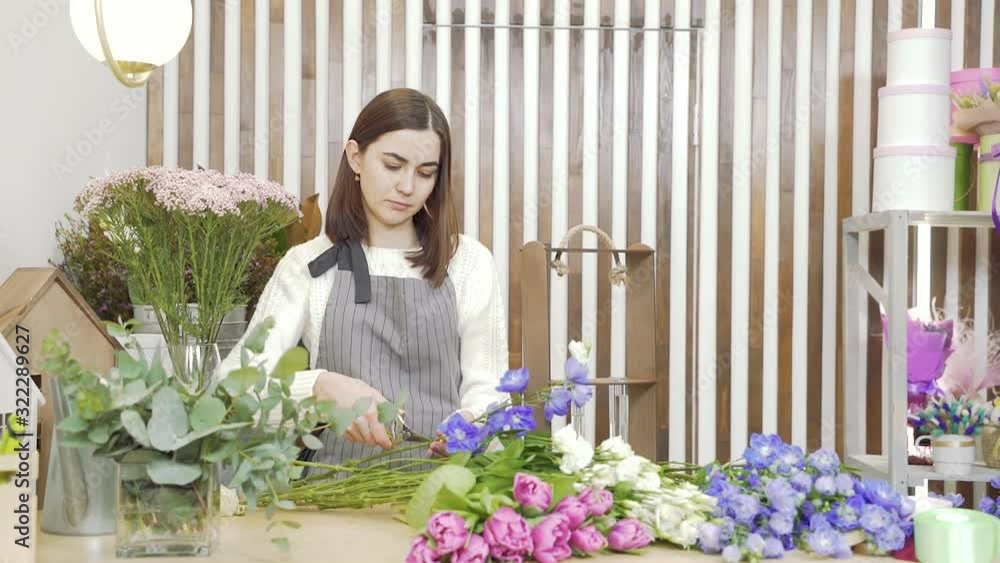 Young woman florist preparing flowers for creating a bouquet or composition, inspecting and pruning them in the workshop
