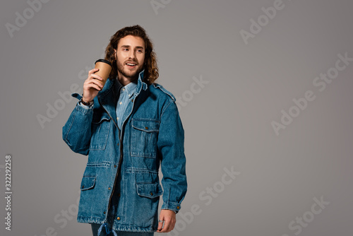 smiling and stylish man in denim jacket holding paper cup isolated on grey
