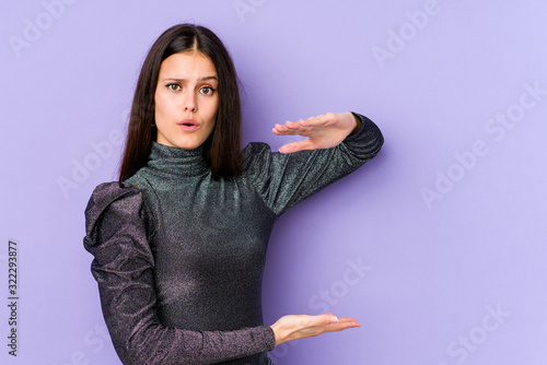 Young caucasian woman isolated on purple background holding something with both hands, product presentation. © Asier