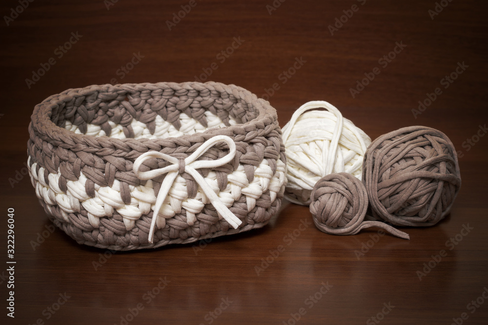 Fototapeta premium Knitted basket of handmade knitted yarn on a wooden background. Around the basket are balls of knitted yarn in white and brown.