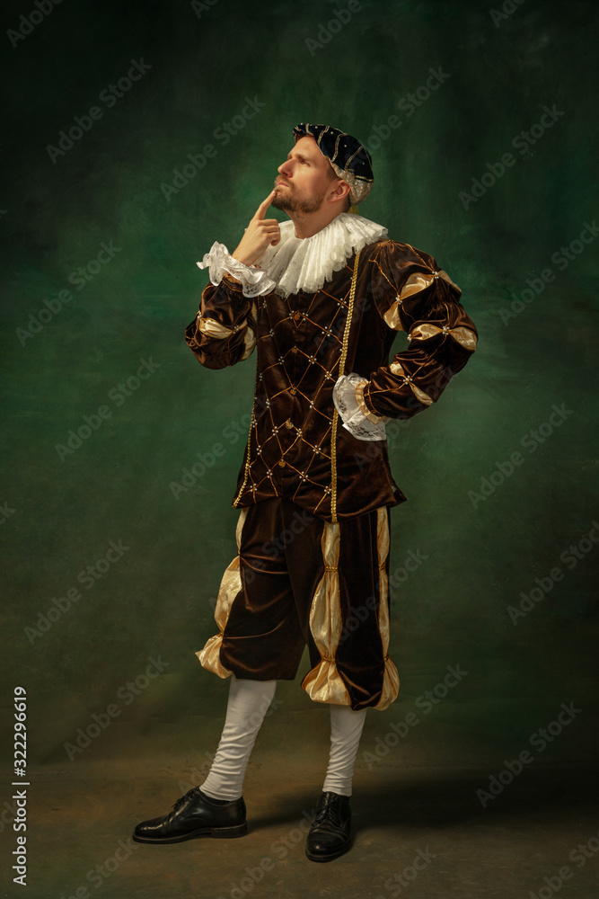 Posing thoughtful. Portrait of medieval young man in vintage clothing standing on dark background. Male model as a duke, prince, royal person. Concept of comparison of eras, modern, fashion.