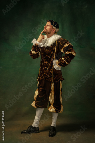 Posing thoughtful. Portrait of medieval young man in vintage clothing standing on dark background. Male model as a duke, prince, royal person. Concept of comparison of eras, modern, fashion. photo