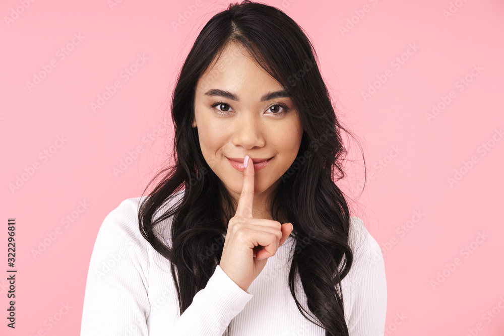 Image of young brunette asian woman smiling and holding finger on lips Image of young brunette asian woman smiling and holding finger on lips