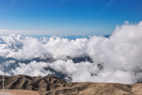 Aerial view of amazing scenic sunny landscape. Tops of high rocky mountains and white soft fluffy clouds flying around in blue sky covering peaks of mounts.