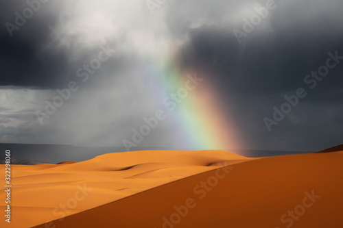 Sahara desert sand dunes with rainbow against dark, cloudy, rainy sky.