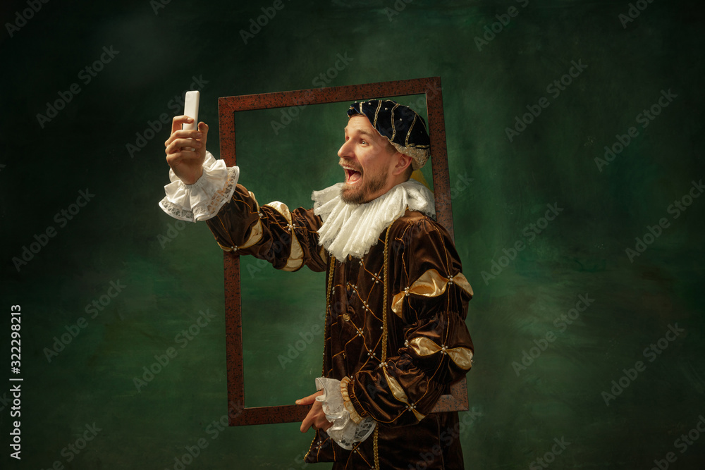 Selfie time. Portrait of medieval young man in vintage clothing with wooden frame on dark background. Male model as a duke, prince, royal person. Concept of comparison of eras, modern, fashion.