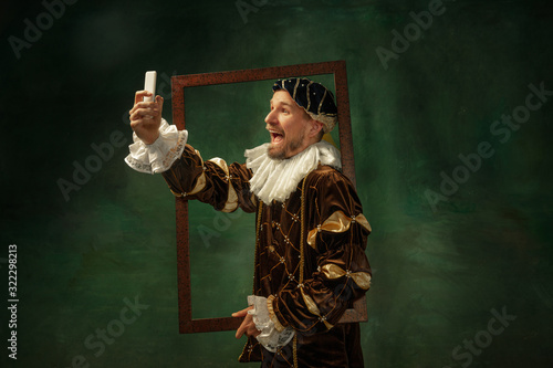 Selfie time. Portrait of medieval young man in vintage clothing with wooden frame on dark background. Male model as a duke, prince, royal person. Concept of comparison of eras, modern, fashion.