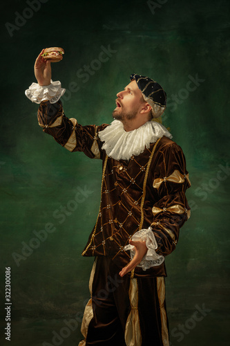 Shocking food. Portrait of medieval young man in vintage clothing with wooden frame on dark background. Male model as a duke, prince, royal person. Concept of comparison of eras, modern, fashion.