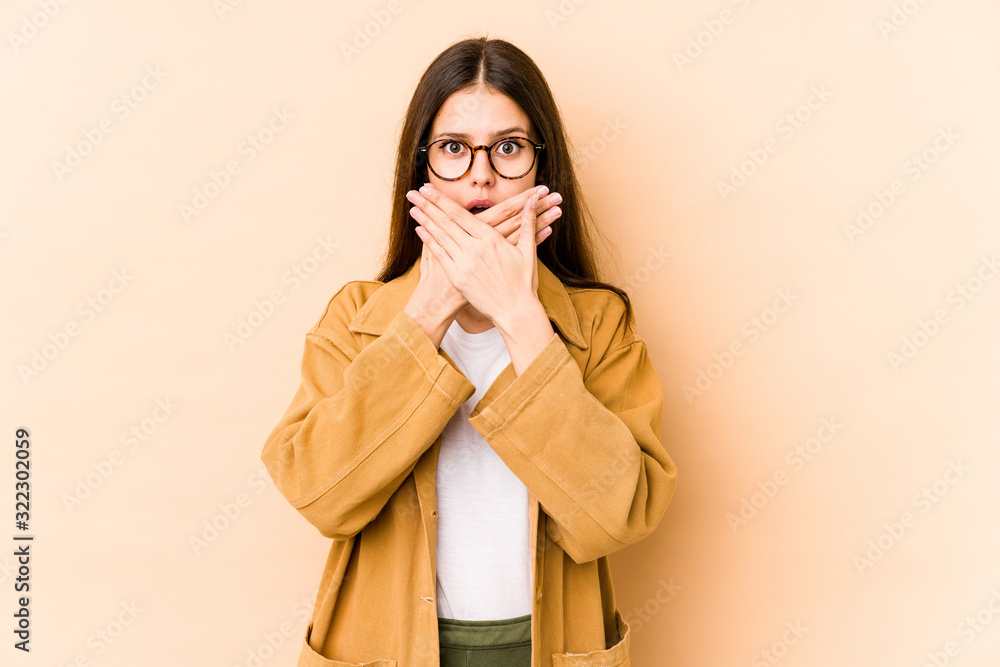 Young caucasian woman isolated on beige background shocked covering mouth with hands. Young caucasian woman isolated on beige background shocked covering mouth with hands.