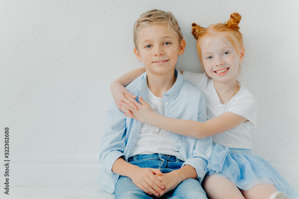 Friendly small girl and boy hug and sit on floor against white ...