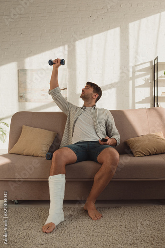 Handsome man with leg in plaster bandage training with dumbbell and holding remote controller on couch © LIGHTFIELD STUDIOS