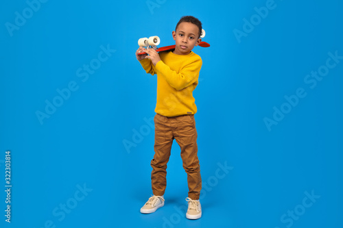 Funny little African-American boy with a skateboard smiles and looks away, standing on a blue background. Concept of activity and happy childhood.