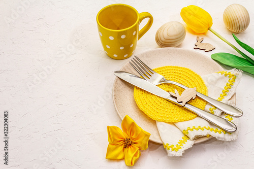 Easter table setting on textured white putty background. Spring holiday card template. Cutlery, knitted napkin, egg, bunny, tulip, polka dot cup