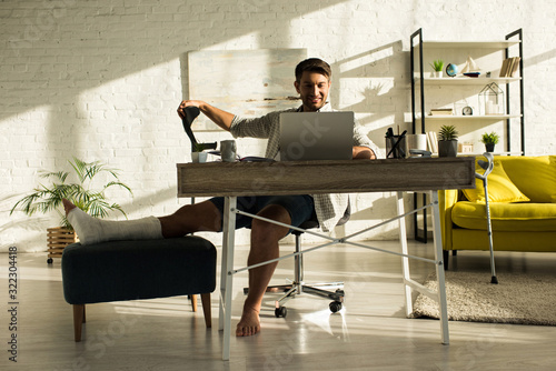 Smiling freelancer with broken leg working at laptop in living room
