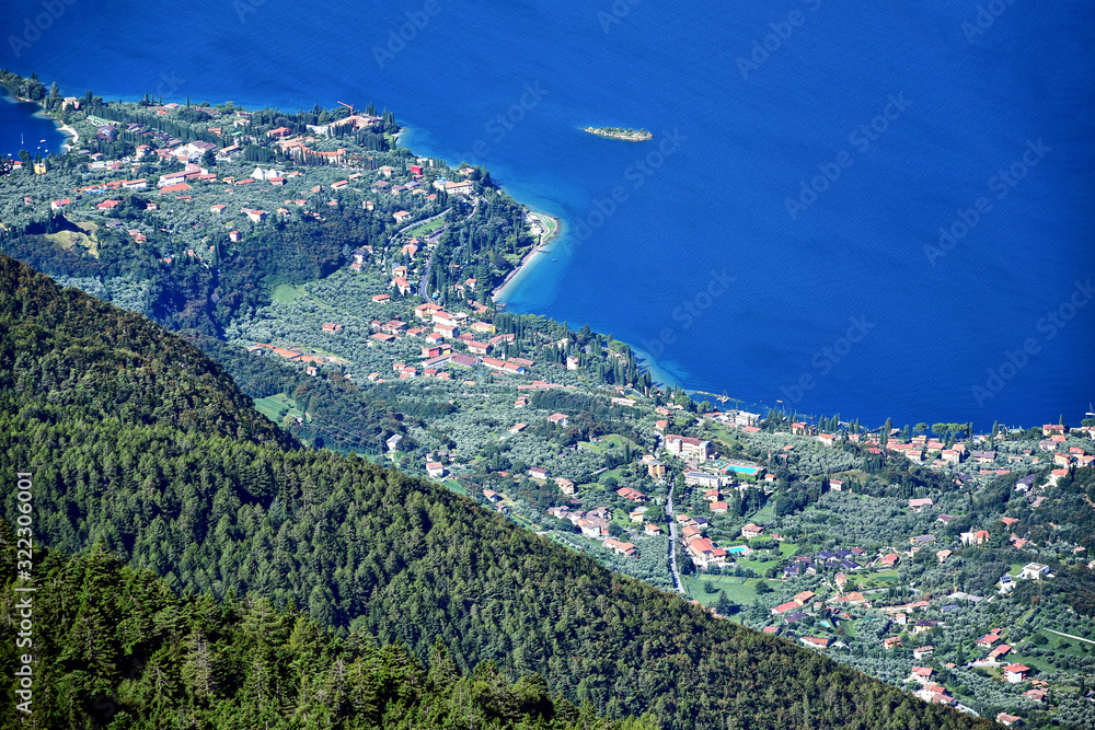 Monte Baldo, Italy - mountains covered with greenery and trees, aerial view of the city of Malcesine and the blue Lake Garda, in the summer afternoon.