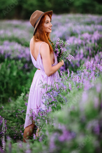 Red-haired girl in a hat lies in the grass with purple flowers. Young woman smile in nature. lady walks on a lavender field. © alex_shifer
