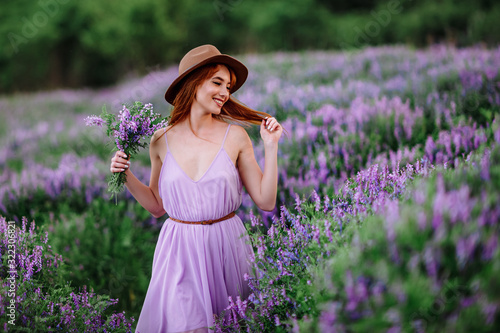 Red-haired girl in a hat lies in the grass with purple flowers. Young woman smile in nature. lady walks on a lavender field.