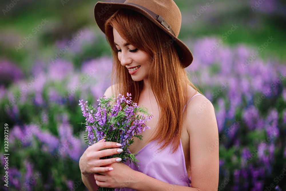 Red-haired girl in a hat lies in the grass with purple flowers. Young woman smile in nature. lady walks on a lavender field.