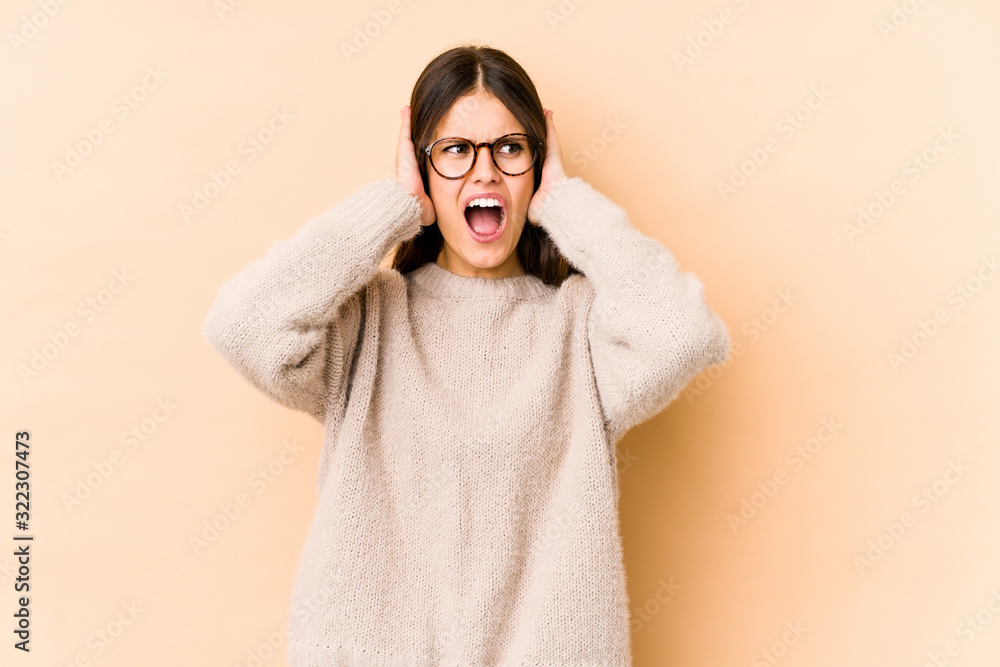 Young caucasian woman isolated on beige background covering ears with hands trying not to hear too loud sound. Young caucasian woman isolated on beige background covering ears with hands trying not to hear too loud sound.