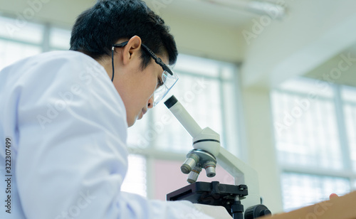 close up rear view of young asian scientist looking at microscope to research or record and analyze about scientific experiment result at laboratory for healthcare and pharmaceutical concept