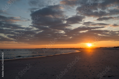 The sun sets behind some clouds along the coastline of a sandy beach with a colorful sky.