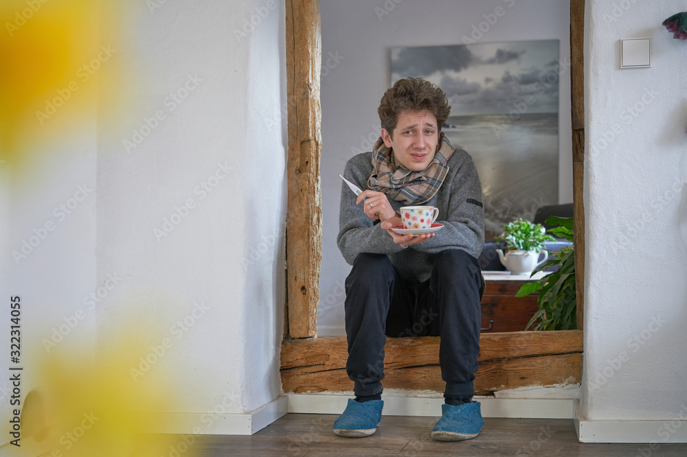 Young curly man feeling roughly, sitting with a cup of tea and a thermometer and a scarf in a door frame, lense flare