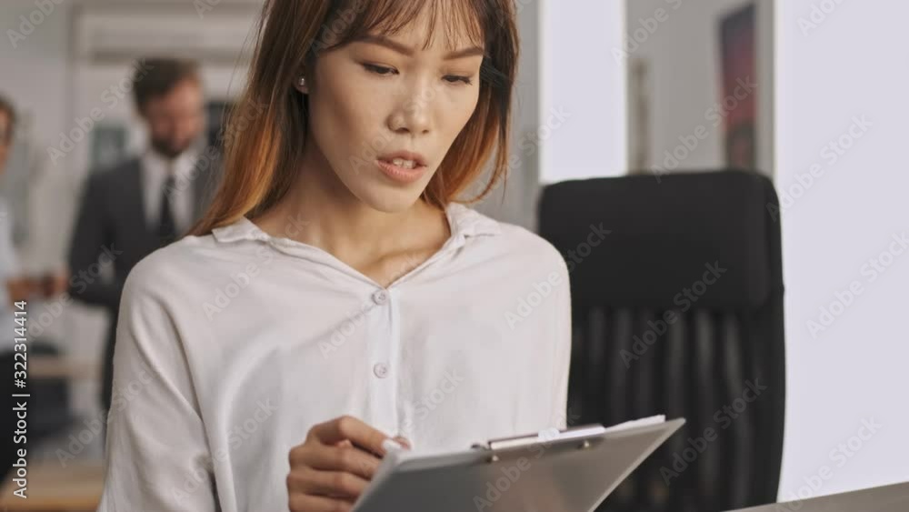 Concentrated asian businesswoman holding clipboard and reading documents while standing in office Concentrated asian businesswoman holding clipboard and reading documents while standing in office