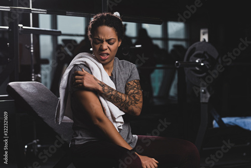injured african american woman with tattoo touching hand while suffering from pain in gym