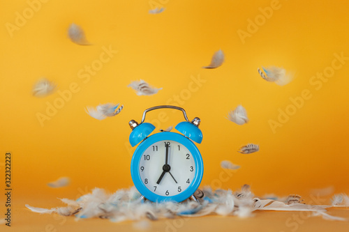 Soft fluffy flying feathers and blue alarm clock on yellow background. photo