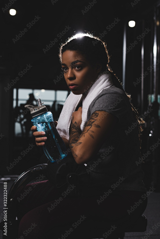 Naklejka premium young african american girl with tattoo holding sports bottle in gym