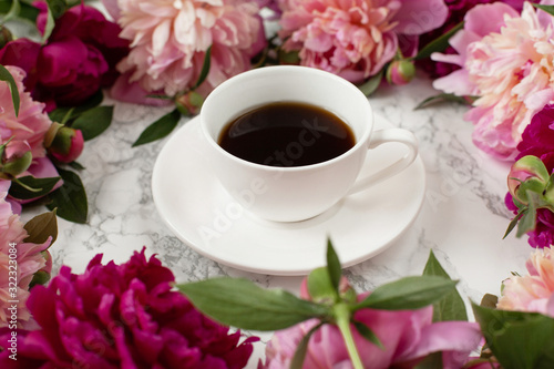 White cup with hot coffee or tea on a white marble background. Pink and red peonies around the mug. Beautiful still live with drink and fresh flowers, close up. International womans day concept