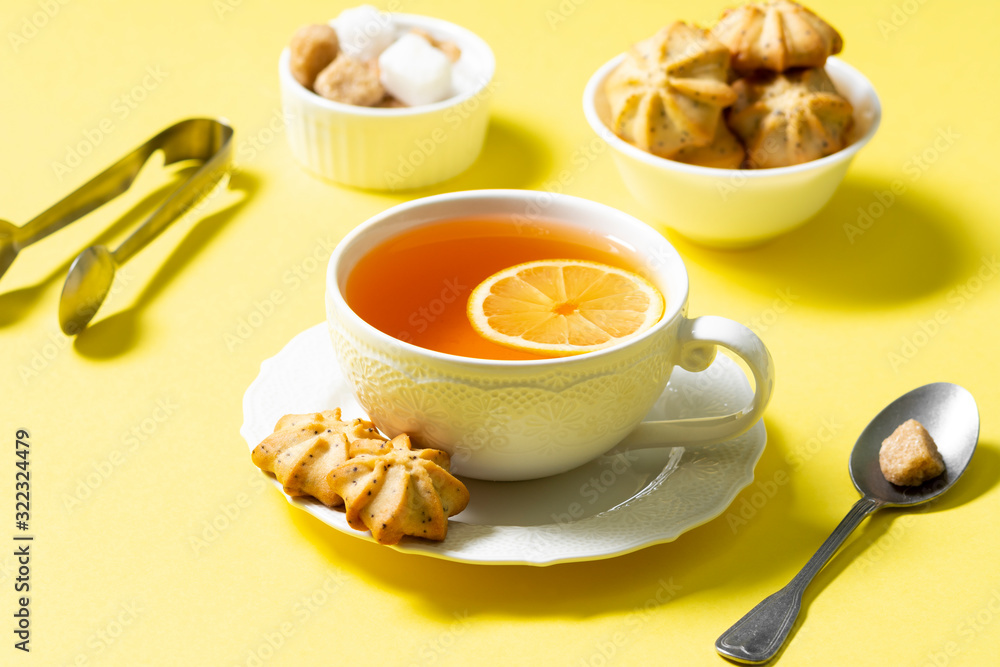 Tea with lemon in a white Cup and lemon cookies with poppy seeds in a white ceramic bowl on a yellow background. The concept of Breakfast or snack Tea with lemon in a white Cup and lemon cookies with poppy seeds in a white ceramic bowl on a yellow background. The concept of Breakfast or snack