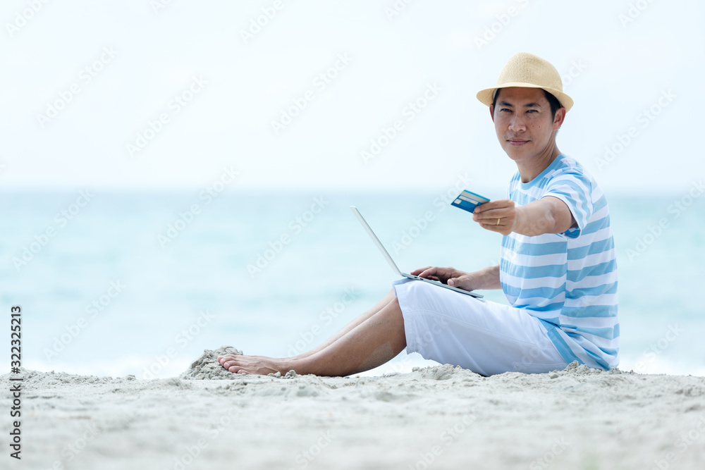 Lifestyle young asian man use credit card on laptop while sitting chill on the beautiful outdoor beach, freelance working social on holiday summer. Summer and Vacation Concept