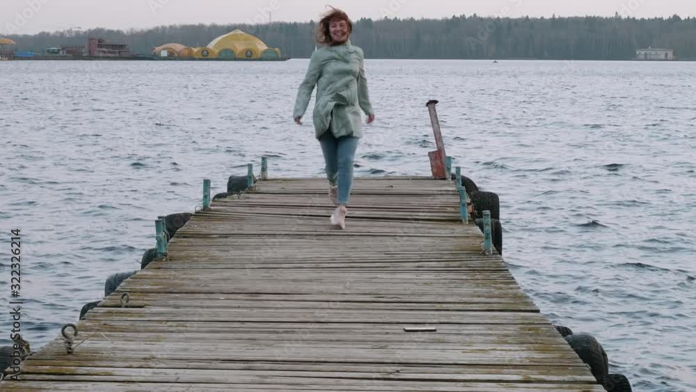 Glad woman in blue coat and jeans and pink sport shoes runs on old wooden pier. Happy woman. Glad woman in blue coat and jeans and pink sport shoes runs on old wooden pier. Happy woman.