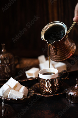 Traditional turkish coffee and turkish delight in traditional oriental copper serving set. Coffee pouring from cezve into a cup. Dark vintage background photo