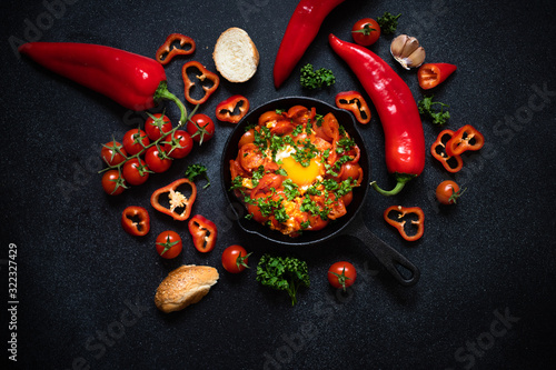 Shakshouka eggs poached in a sauce of tomatoes, vegetables, spices and herbs in iron pan. Bread, tomatoes, red peppers, garlic, parsley. Traditional breakfast. Black background, top view, copy space photo