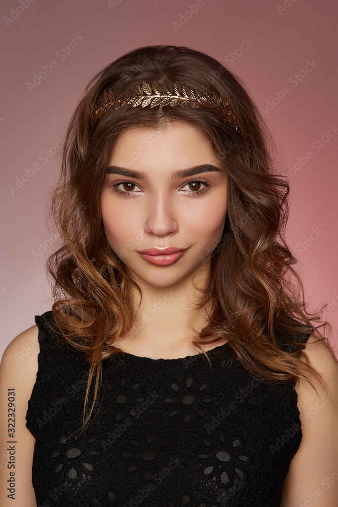 A brown-haired European girl in a black lacy top is posing on the pink background. The girl's wavy hair is fixed with a metal headband made as a crown of silver embossed leaves. A brown-haired European girl in a black lacy top is posing on the pink background. The girl's wavy hair is fixed with a metal headband made as a crown of silver embossed leaves.