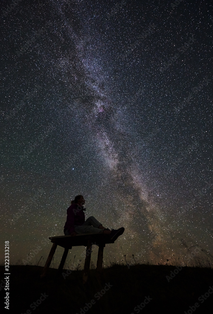 Dark silhouette of female hiker resting at summer night camping in the mountains. Young woman sitting on a bench, enjoying view of night sky full of stars and Milky way. Astrophotography Dark silhouette of female hiker resting at summer night camping in the mountains. Young woman sitting on a bench, enjoying view of night sky full of stars and Milky way. Astrophotography