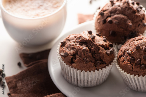 close up view of fresh chocolate muffins on white plate and brown napkin near cappuccino on marble surface