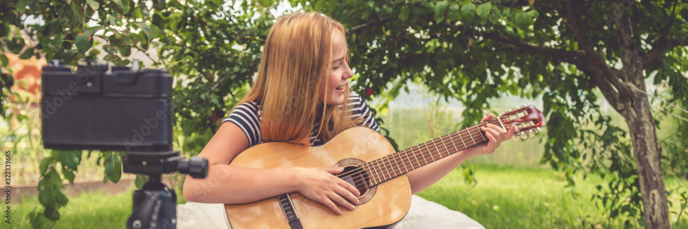 Fototapeta premium Teen girl shoots a blog on music and learning online guitar playing in nature in front of the camera, the idea of youth hobbies