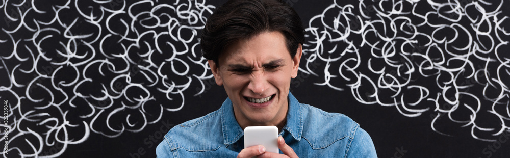 panoramic shot of worried young man using smartphone, with steam from ears drawing on chalkboard panoramic shot of worried young man using smartphone, with steam from ears drawing on chalkboard