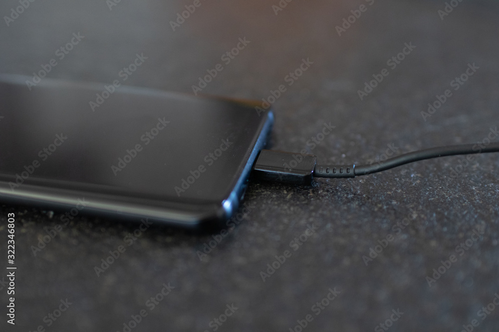 Smartphone charging, detail of usb c cable connector. Charging phone connected with wire on an elegant black stone table. Smart work smartphone in charging battery. Plugged device to power source Smartphone charging, detail of usb c cable connector. Charging phone connected with wire on an elegant black stone table. Smart work smartphone in charging battery. Plugged device to power source