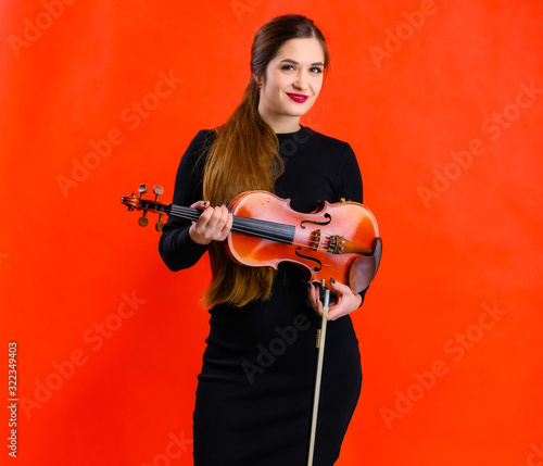 Portrait of a pretty brunette musician girl with a smile in a black dress on a red background holds a violin in her hands