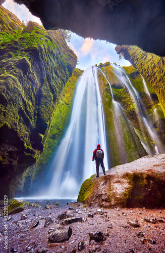 Unique incredible Gljufrabui waterfall in cave, hidden place. One of the most beautiful waterfalls on the Iceland. Man hiker in red jacket standing on stone and looks at flow of falling water Postcard