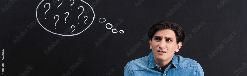 panoramic shot of confused young man with question marks in thought bubble on blackboard panoramic shot of confused young man with question marks in thought bubble on blackboard