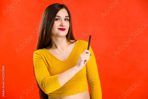 nice brunette girl with long hair with a smile with a pen in her hands in a yellow sweater is happy over a red background smiling and showing positive emotions