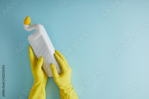 cropped view of housekeeper in yellow rubber gloves holding bottle with toilet cleaner on blue