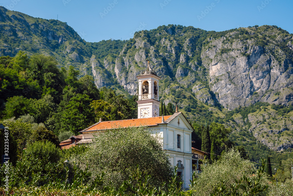 An old italian Catholic church (Chiesa dei Santi Nabore e Felice) surrounded by lush green vegetation, at the foot of a high rocky mountain, Griante, Lake Como, Italy.